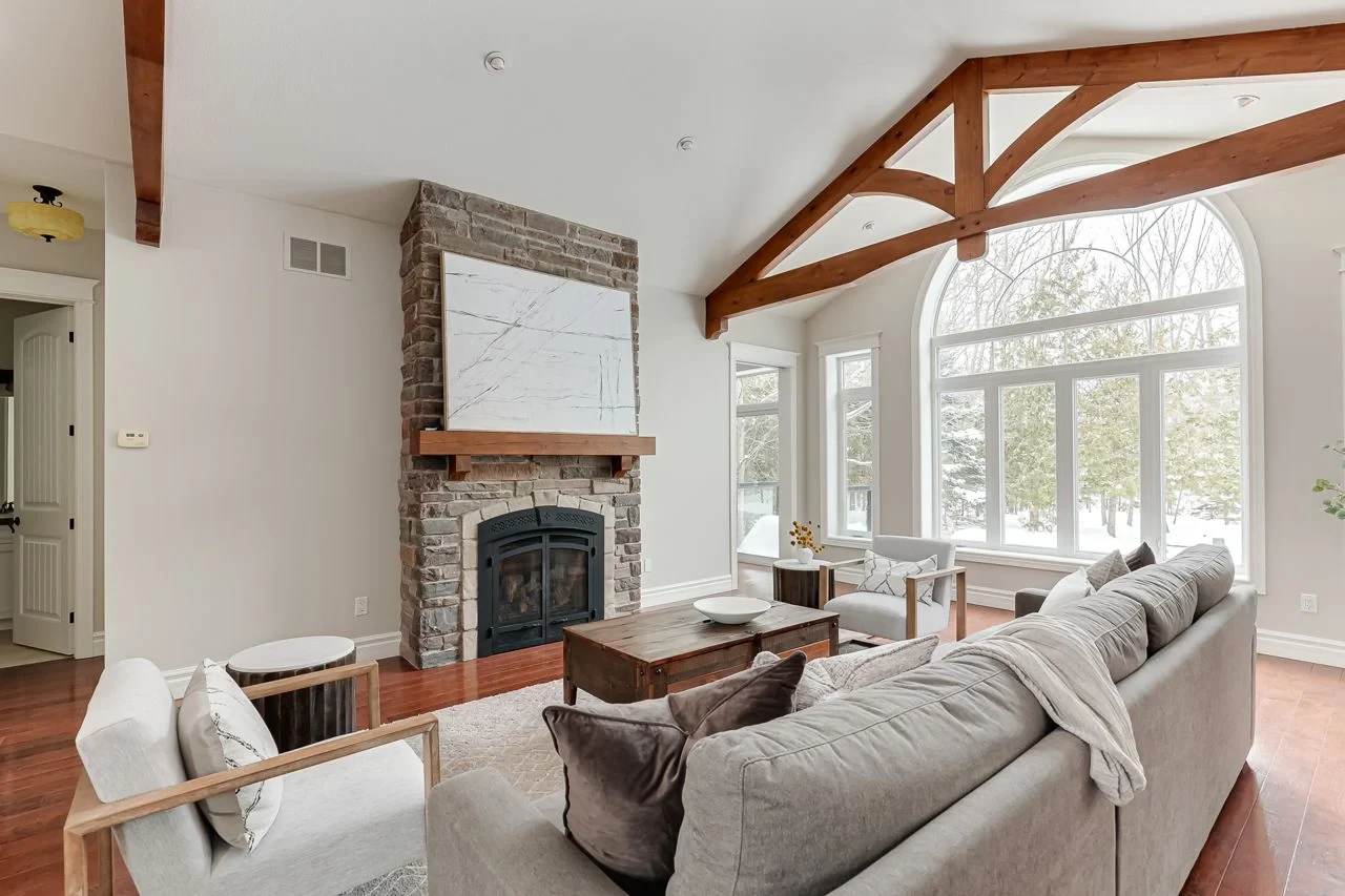 Cozy four-season cottage living room with stone fireplace and snow-covered trees visible through large windows in Muskoka