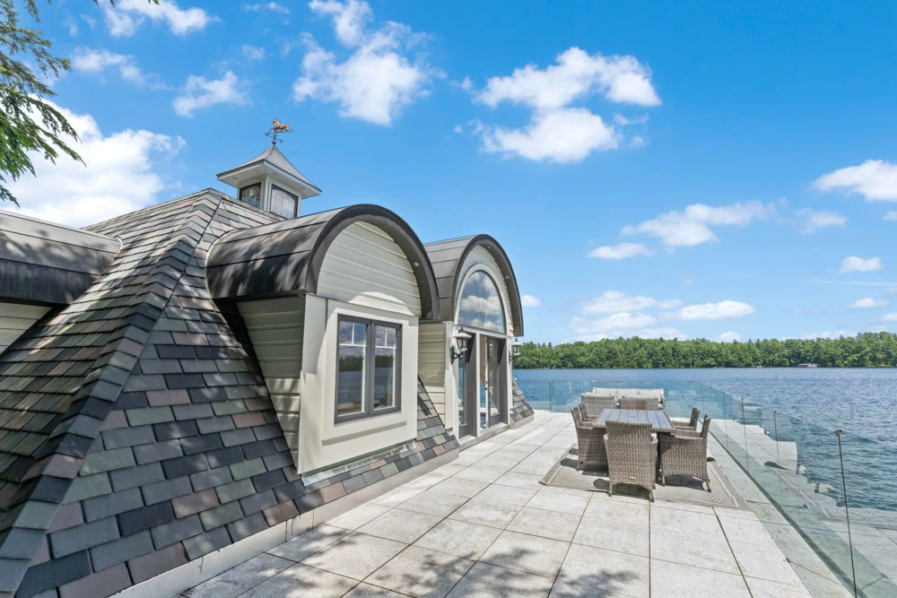 Ontario boathouse rooftop deck with curved shingle roof, arched dormer window, outdoor dining furniture, and calm lake views in Seguin