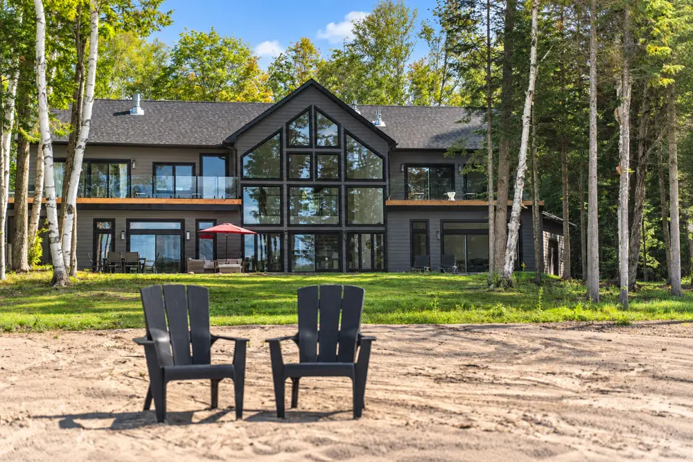 Exterior of a modern Haliburton waterfront cottage with floor-to-ceiling windows, dark cladding, and Muskoka chairs on a sandy lakefront area